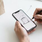 Tournage d’un Déballage Produit – merci e-commerce Un entrepreneur filme un déballage au smartphone, avec un sac et un colis sur son bureau.